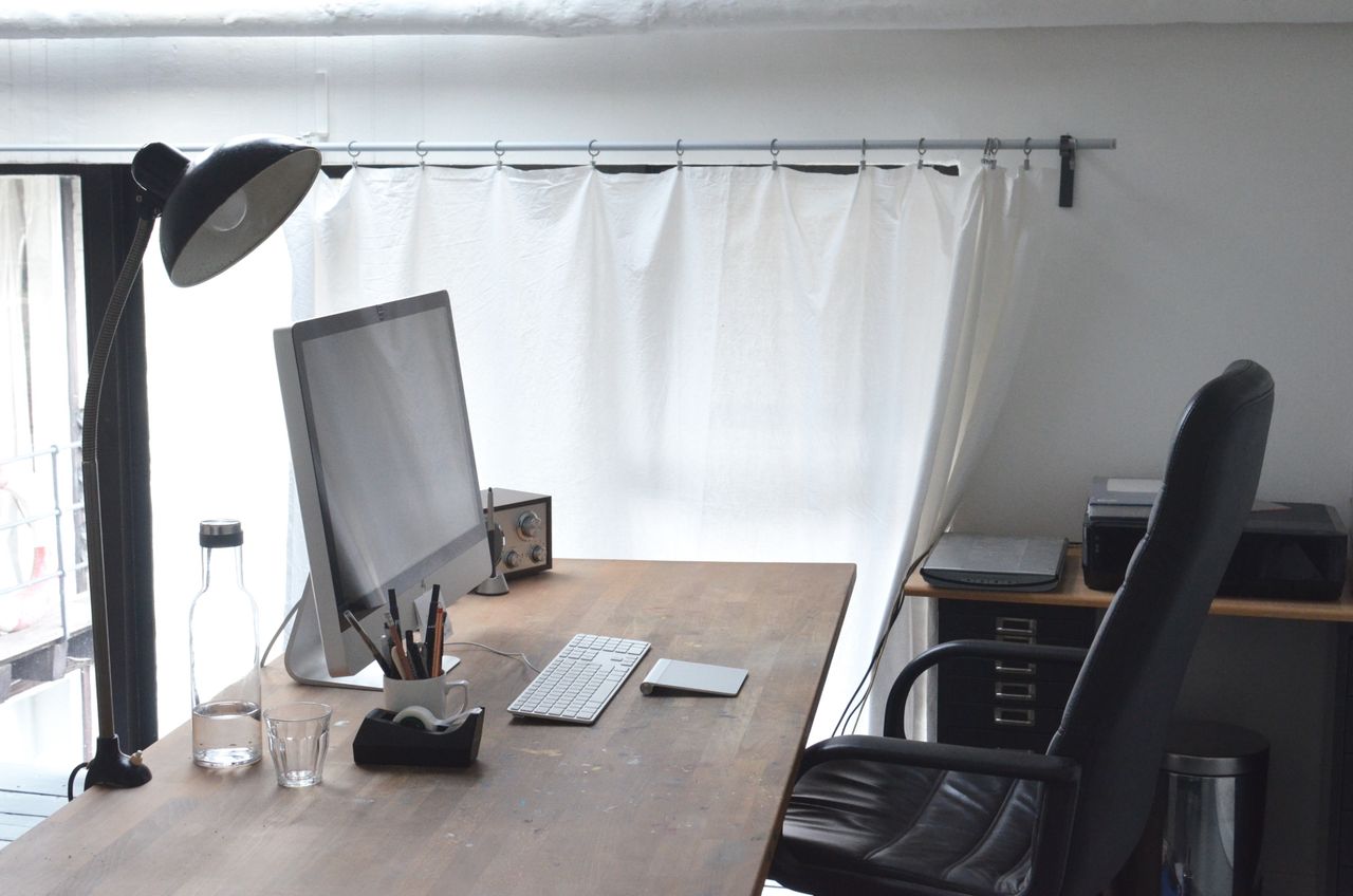 Modern computer with stationery and lamp arranged on wooden table near comfortable leather chair in office